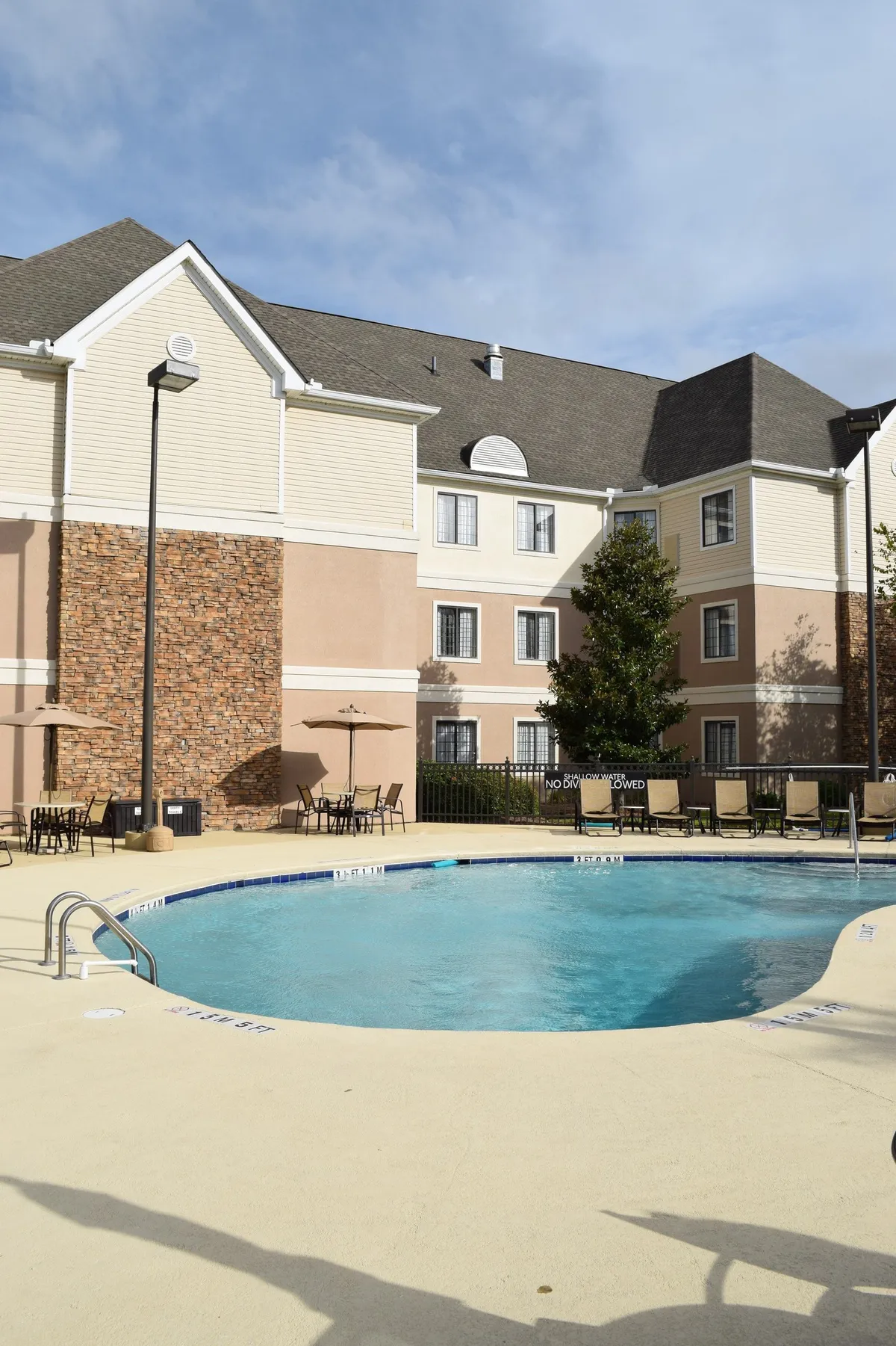 This exterior shot presents a different angle of the swimming pool, focusing on the calm water, inviting lounge chairs, and a glimpse of the building facade in the background. The ambiance is peaceful and inviting, with shaded areas under umbrellas perfect for relaxing or social gatherings. The scene hints at the comfort and convenience of on-site amenities accessible right outside the residences.