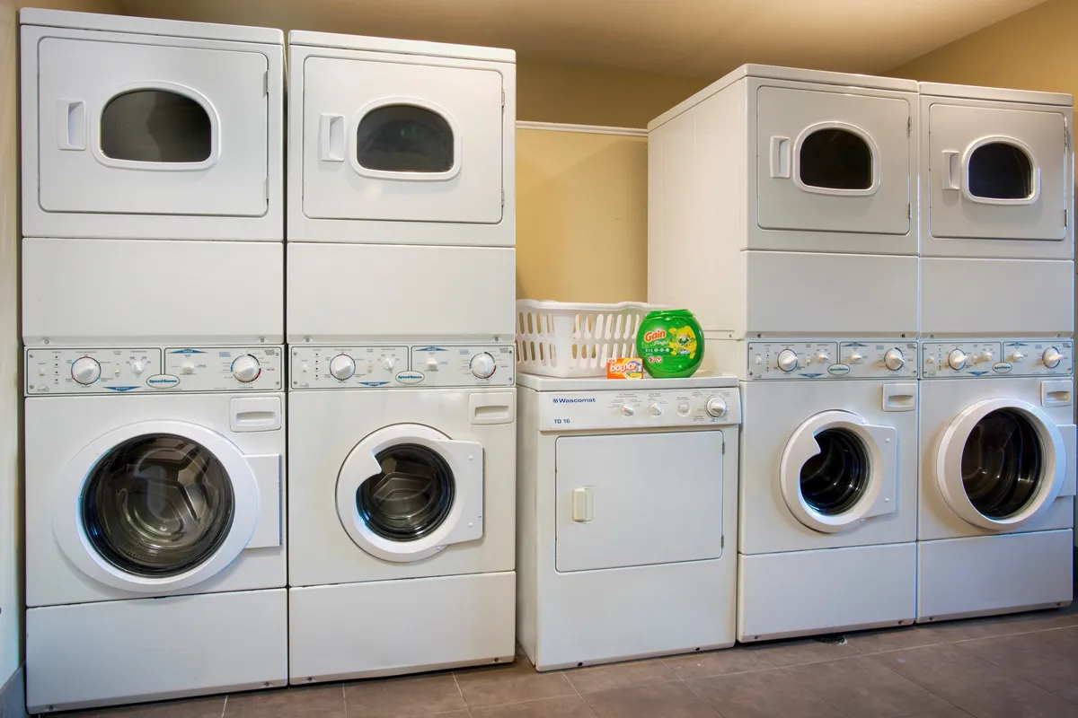 The photo shows a practical laundry area with modern washer and dryer units, a laundry basket, and cleaning supplies neatly organized. The space is clean and functional, illustrating the convenience of on-site laundry facilities.
