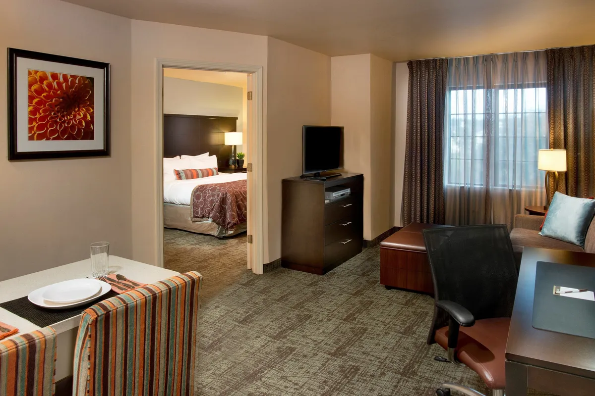 The image showcases a bright, inviting open-plan living and dining area with a table and chairs set for shared meals, a large window allowing in natural light, a comfortable seating area, a television atop a dresser, and a doorway leading to the cozy bedroom. The space feels both welcoming and practical, ideal for relaxing after a day at the beach or catching up on work.