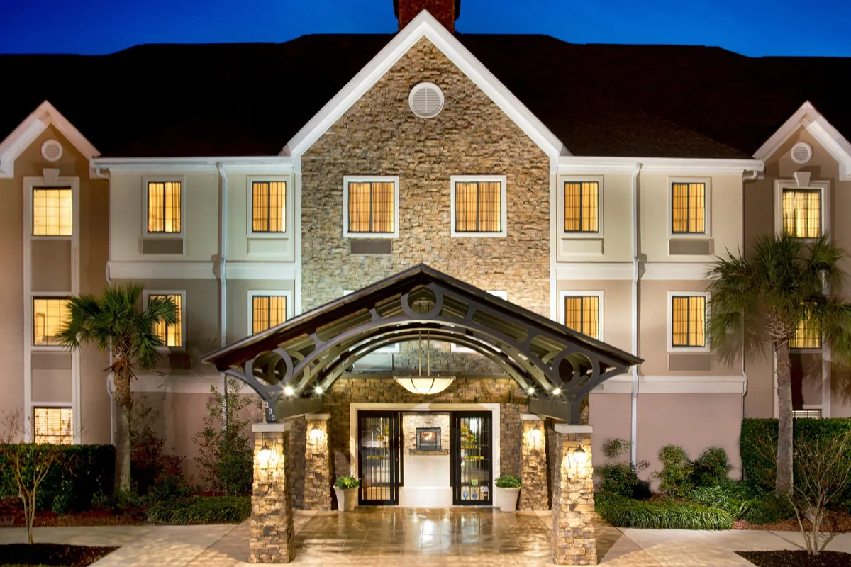 This image showcases the welcoming exterior of Staybridge Suites, featuring the entrance canopy, a charming stone facade, warmly lit windows, and guests engaging in check-in or gathering with friends. The vibrant yet relaxed atmosphere suggests easy arrivals and a great starting point for local adventures.