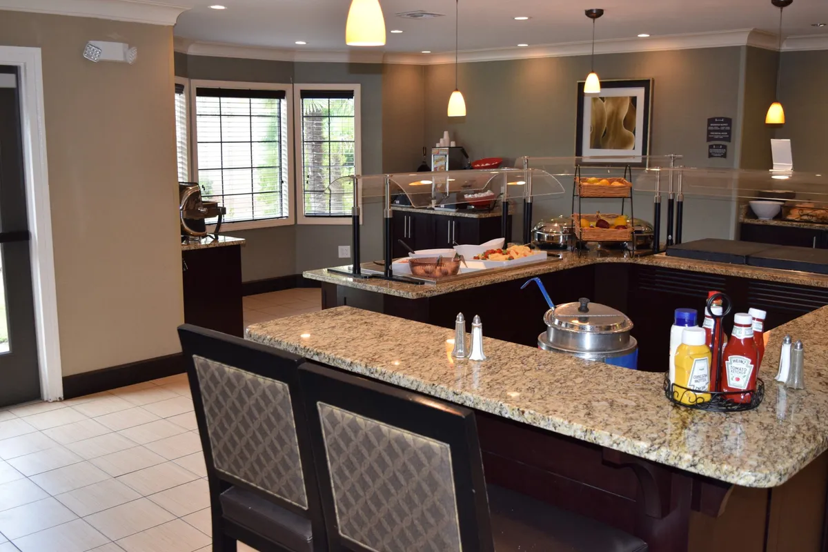The image features a bright and welcoming interior breakfast area with a granite countertop buffet display, bar stools, and large windows letting in natural light. People are shown socializing over breakfast, creating a sense of community and a comfortable atmosphere for guests to start their day.