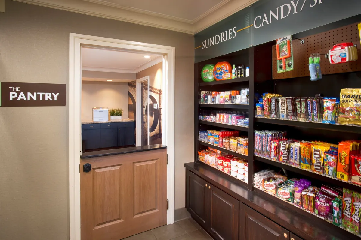 A well-stocked pantry with shelves of snacks and sundries, a wide doorway, and a prominent 'THE PANTRY' sign. It has a casual and accessible feel, inviting guests to grab essentials or treats during their stay.
