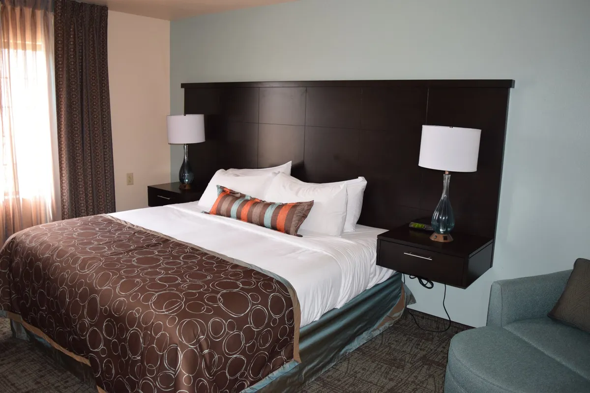 A cozy, well-lit bedroom featuring a neatly made bed with decorative pillows, a nightstand with a lamp, and a comfortable seating area by the window. The soft, neutral decor creates an inviting and restful atmosphere, ideal for winding down after a busy day.